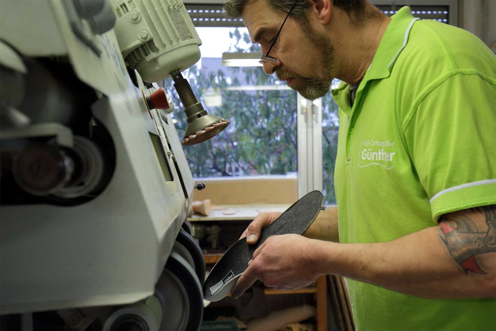 Mann mit Brille und grünem Poloshirt hält eine Schuhsohle vor eine Maschine in einer Werkstatt