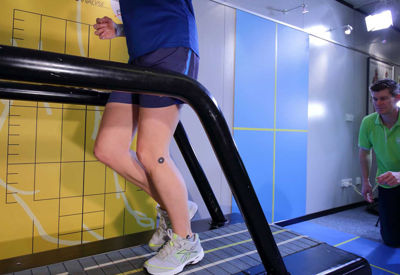 Beine einer Person in Sportschuhen auf einem Laufband, daneben ein Mann in grünem Poloshirt in einem Raum mit gelb-blauen Wänden