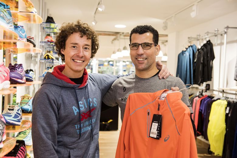 Zwei Männer in Sportbekleidung stehen in einem Sportgeschäft, einer hält eine orangefarbene Jacke mit Preisschild