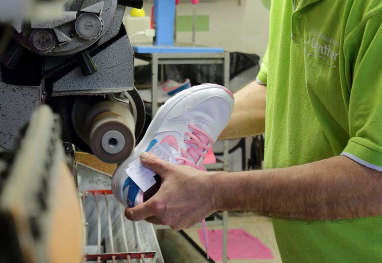 Person hält weißen Sportschuh mit rosa Details vor eine Maschine in einer Werkstatt